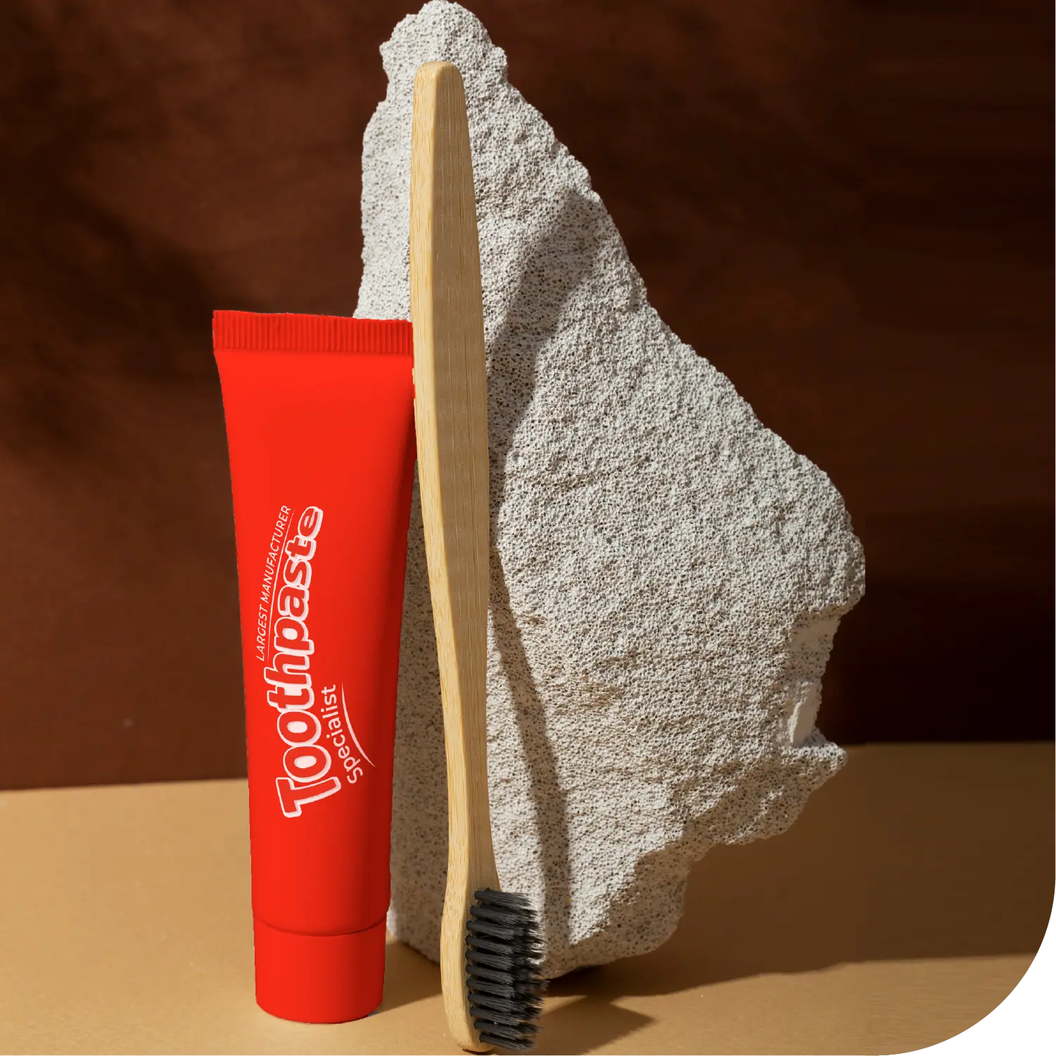 A red toothpaste tube labeled Toothpaste stands upright beside a wooden toothbrush with black bristles, both leaning against a rough-textured pumice stone. The background is a warm brown surface.