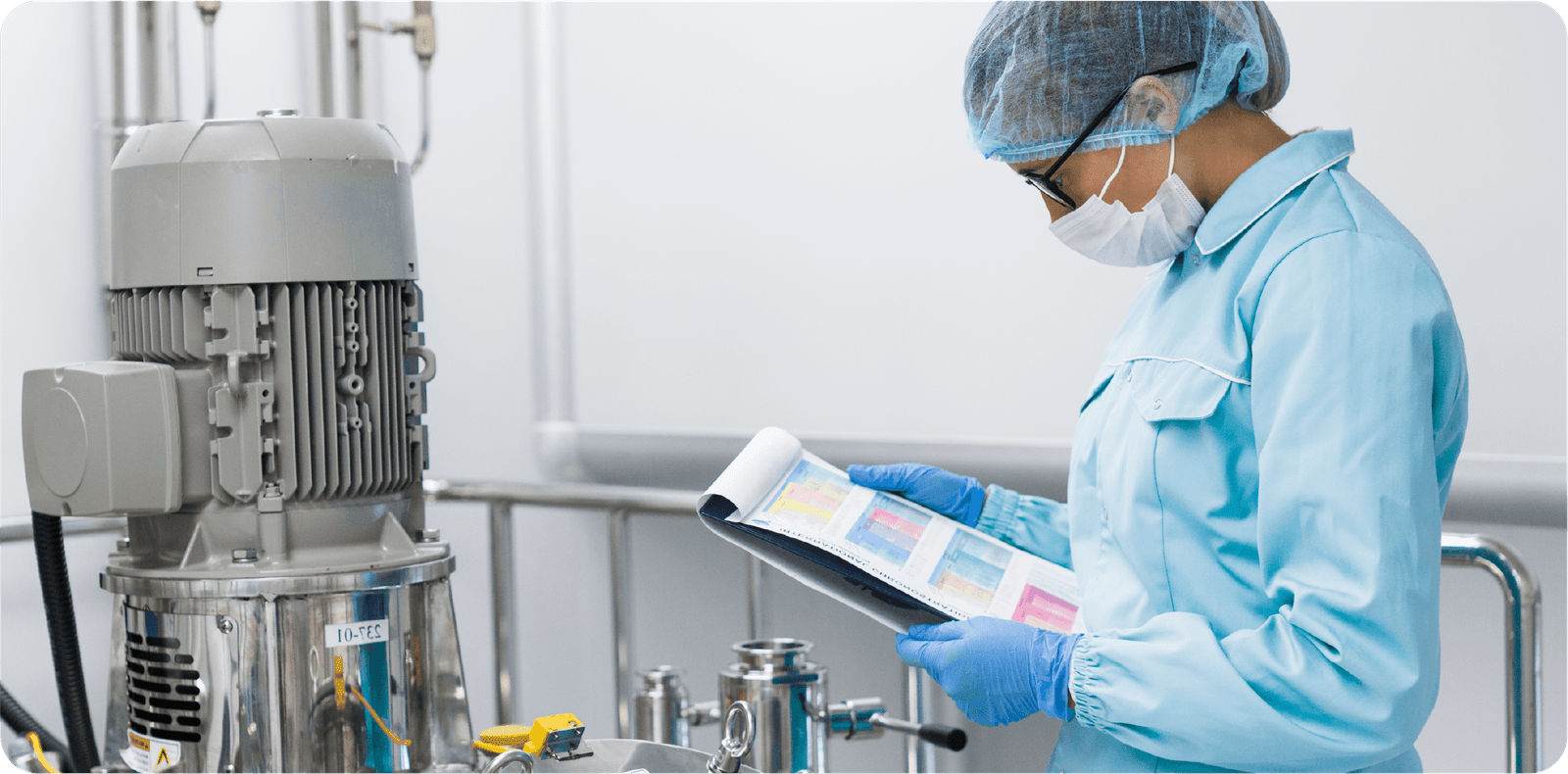 A person in protective clothing, including a mask, hairnet, and gloves, stands in a laboratory or industrial setting. They are reviewing data or charts on a clipboard, next to a large piece of machinery or equipment.