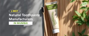 Tube of natural toothpaste and a wooden toothbrush on a bamboo mat. Text reads: 3 Best Natural Toothpaste Manufacturers in Mumbai. A small graphic of a tooth is in the top right corner. Sunlight casts shadows of leaves on the mat.